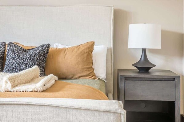Una cama bien hecha con edredón beige y blanco, un plaid gris tejido y una manta blanca. Se observa una mesita de noche con una lámpara blanca y un tocador de color oscuro.