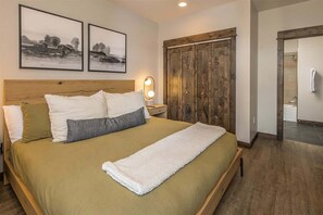 Een slaapkamer met een groot bed, een houten hoofdbord, twee ingelijste foto’s aan de muur, een houten garderobe en een badkamer die door een open deur zichtbaar is.