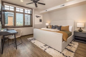 A bedroom with a bed, two bedside tables, a desk, a chair, a ceiling fan, and large windows. - Luxury Two Bedroom Condo - Heart of Town Center (Big Sky)