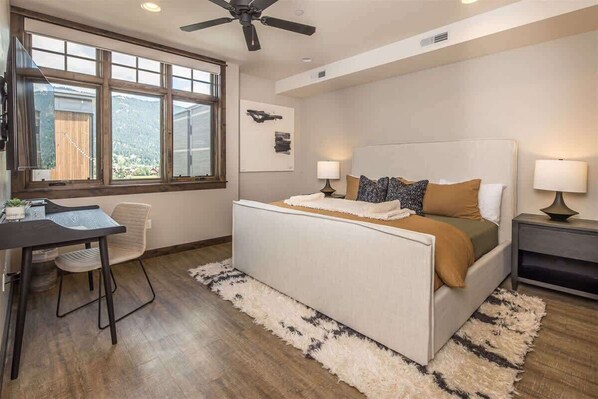 A bedroom with a bed, two bedside tables, a desk, a chair, a ceiling fan, and large windows.