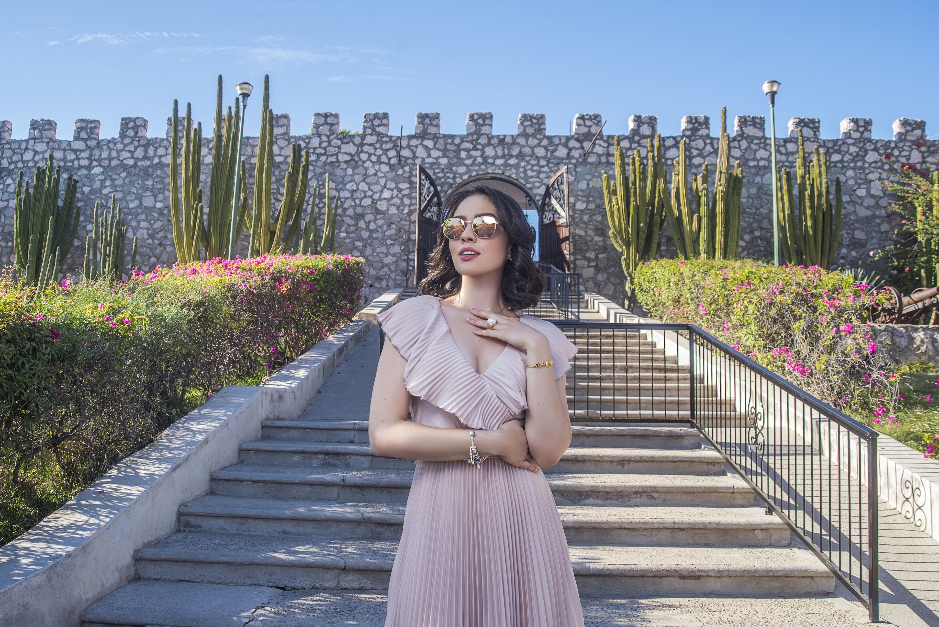 A woman in a dress standing on a stone staircase with a stone wall and cacti in the background.