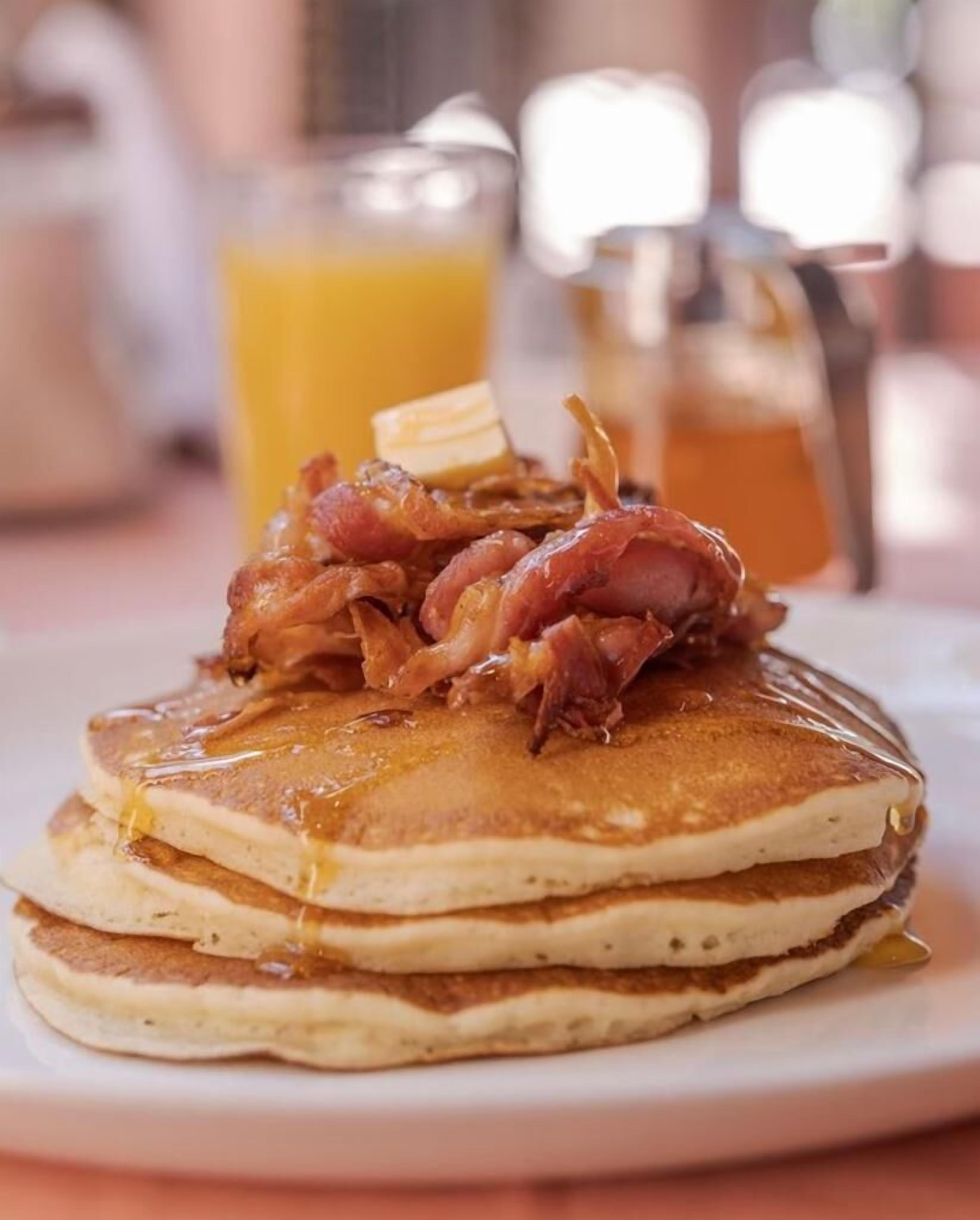 Een stapel pannenkoeken met spek en siroop, met een glas sinaasappelsap en een karaf op de achtergrond.