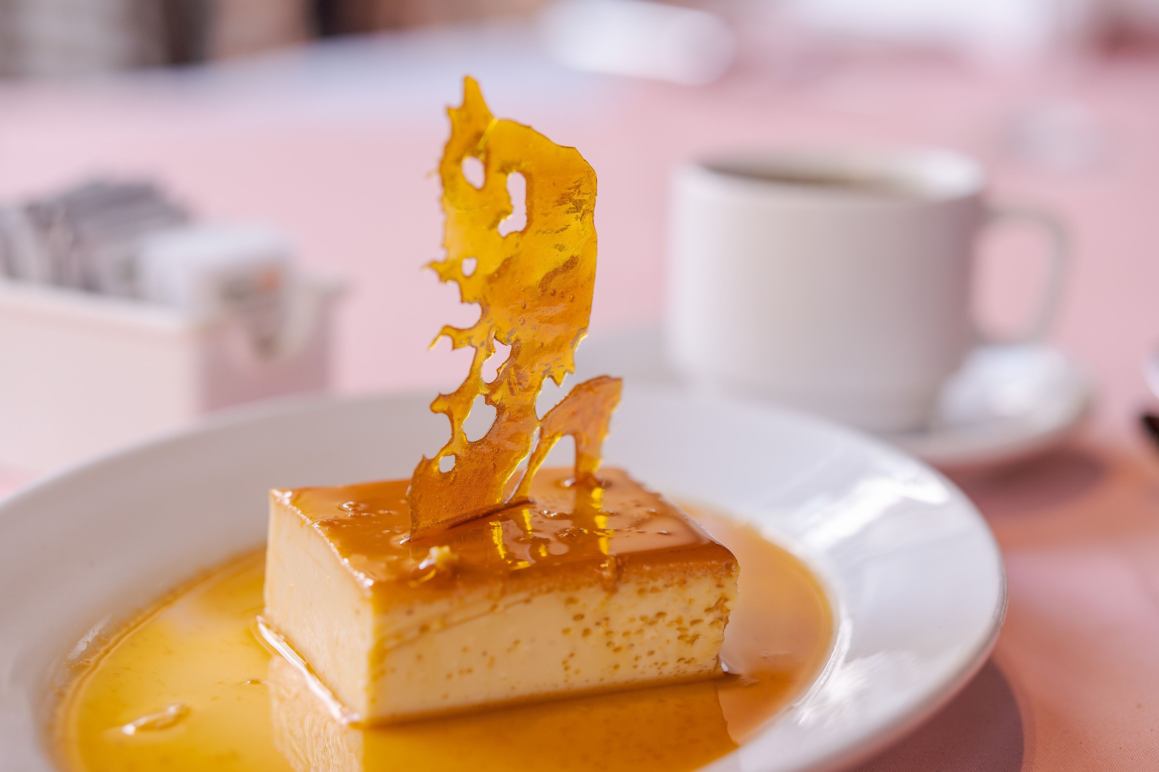 A dessert on a plate with a caramelized topping, a cup of coffee, and a pink tablecloth.