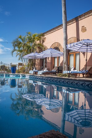A pool area with lounge chairs and umbrellas, palm trees, and a building with arched windows.