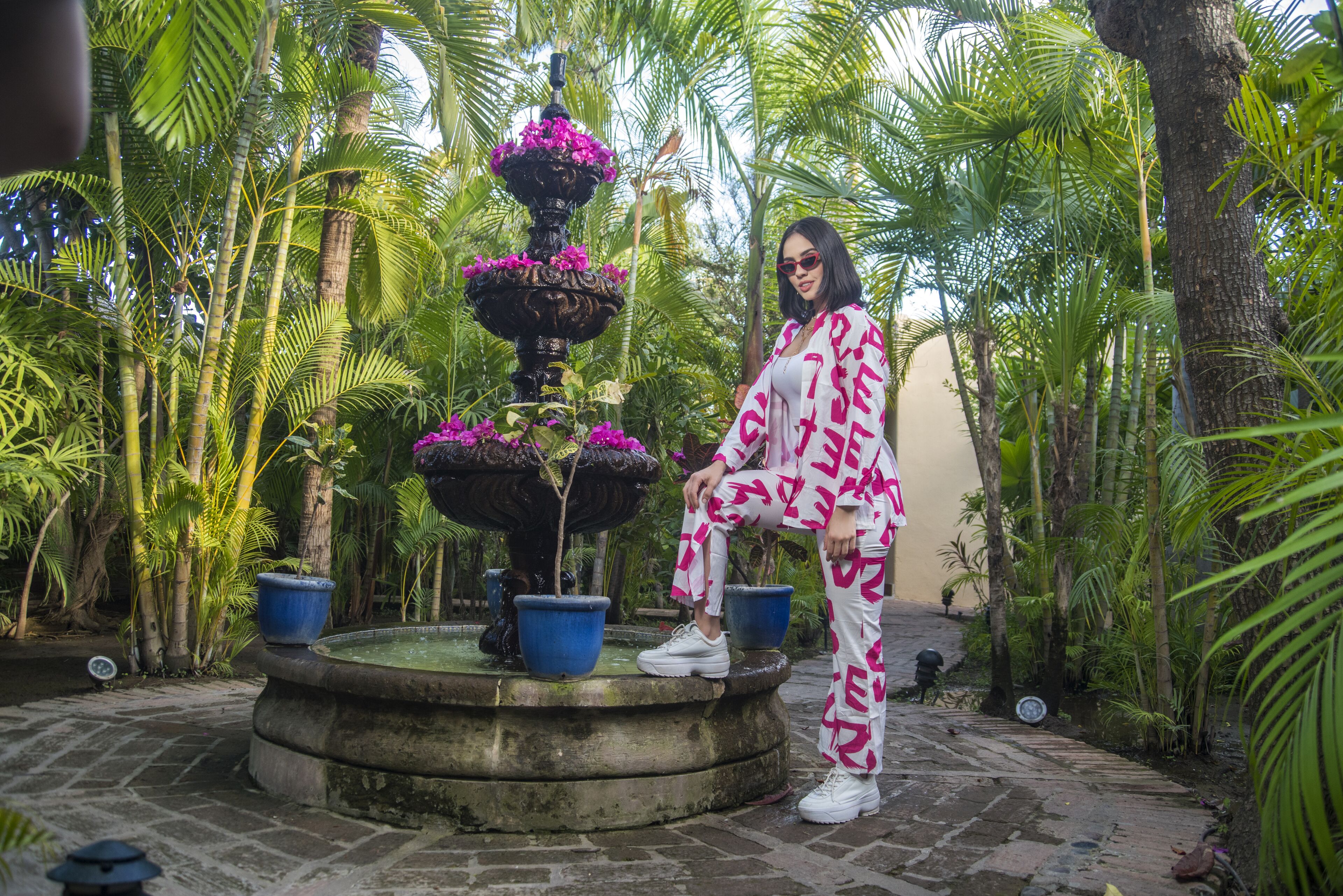 Uma pessoa sentada em um banco de pedra em um jardim tropical, com uma fonte e plantas em vasos.
