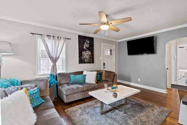 A living room with a ceiling fan, a flat-screen TV, a sofa with patterned cushions, and a coffee table with decorative items.