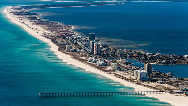 Vista aérea de una zona turística costera con una larga playa de arena, un muelle que se adentra en el océano y un conjunto de edificios a lo largo de la costa.