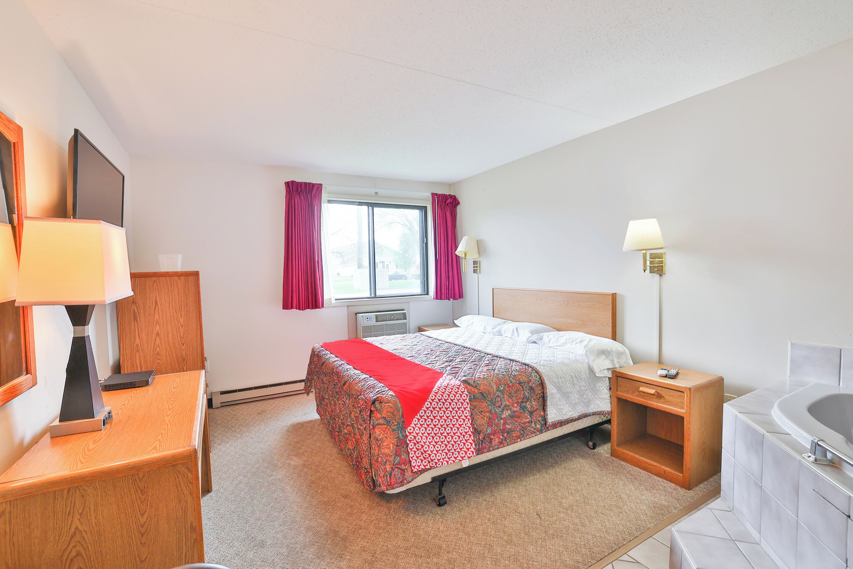 A hotel room with a bed, a desk, a lamp, a window with curtains, a wall-mounted lamp, and a bathtub.
