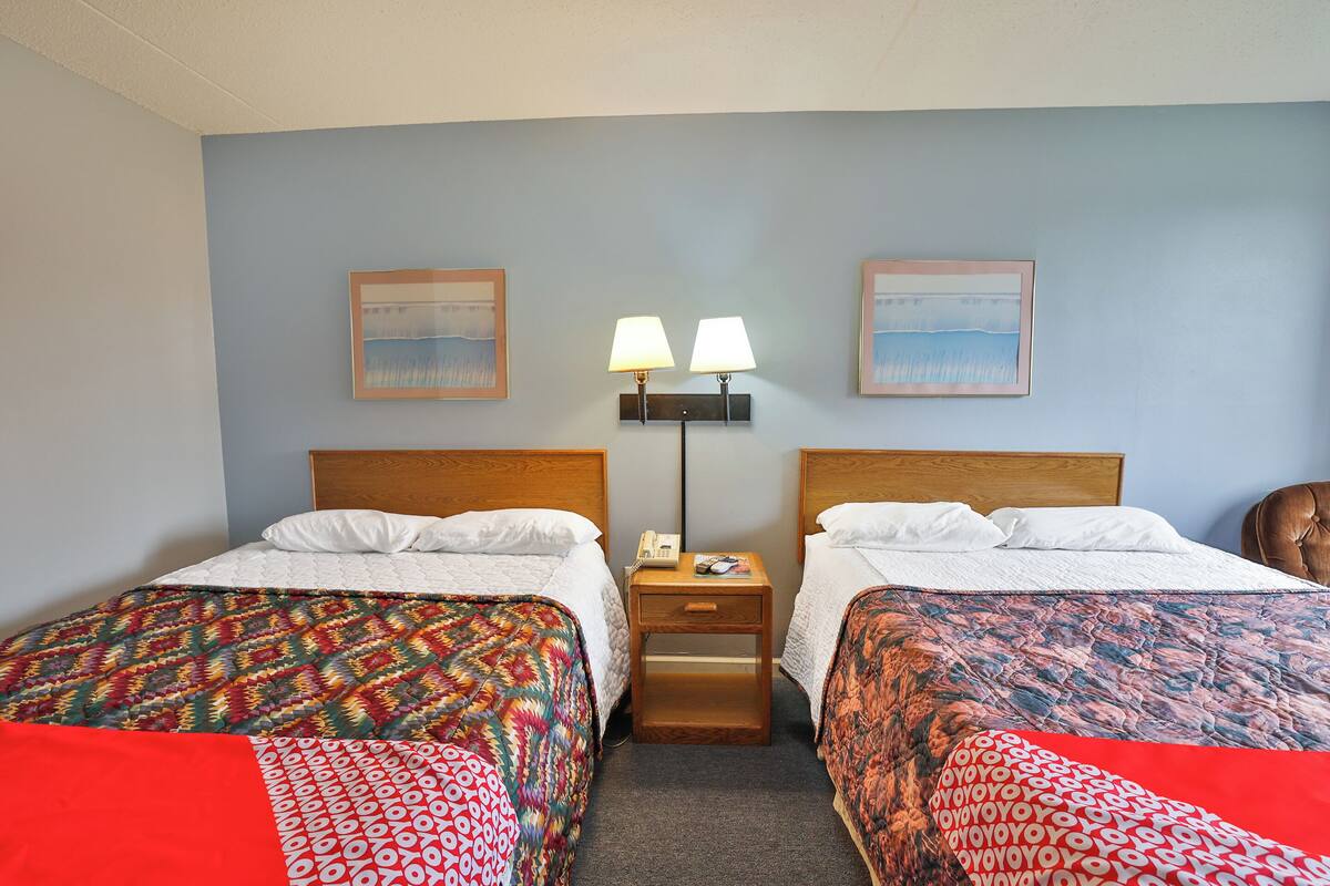 Habitación de hotel con dos camas, una mesita de noche con lámpara y dos cuadros en la pared.