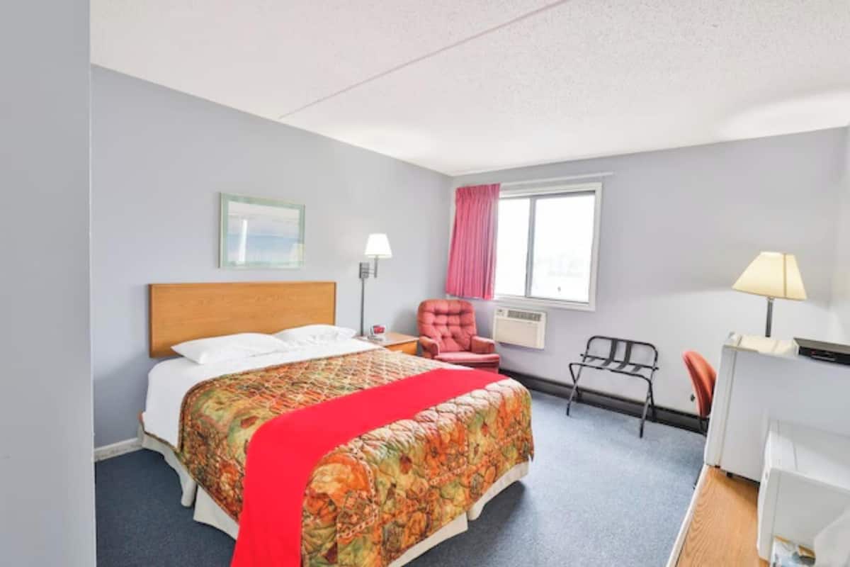 Habitación de hotel con cama, escritorio, silla, ventana con cortinas y lámpara de pared.