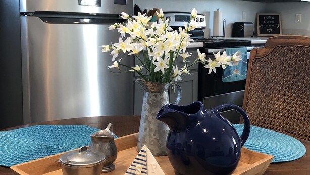 A kitchen with grey cabinets, a stainless steel refrigerator, and a microwave. There is a wooden tray with a vase of flowers, a blue pitcher, and a teapot on the counter. - Grand Lake Barndominium. Close to Wolf Creek & Monkey Island. Lake access! (Afton)