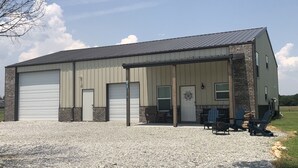 A large industrial building with two garage doors, a covered outdoor seating area with Adirondack chairs, and a gravel driveway. - Grand Lake Barndominium. Close to Wolf Creek & Monkey Island. Lake access! (Afton)