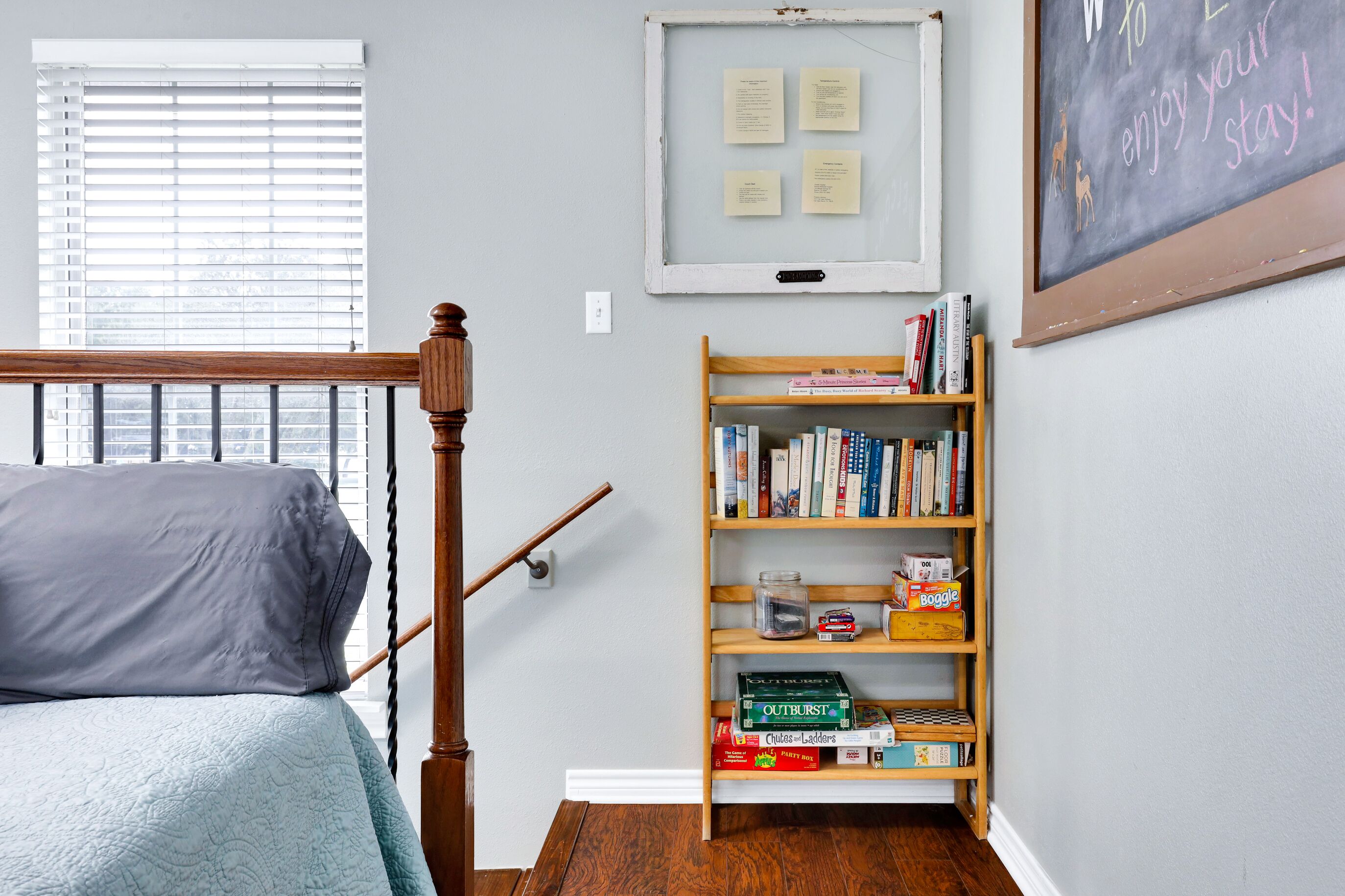A bedroom with a bed, a wooden bookshelf, a window with blinds, and a chalkboard with writing.