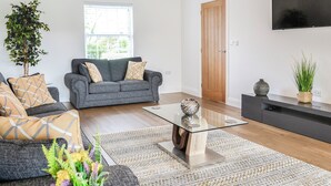 A modern living room with a sofa, a coffee table, a TV, a wooden door, and a potted plant. - 4 bedroom accommodation in Carlisle (Carlisle)
