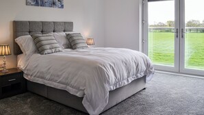 A bedroom with a bed, two bedside tables, a lamp, a window with a view of a grassy area, and two wall-mounted flower paintings. - 4 bedroom accommodation in Carlisle (Carlisle)