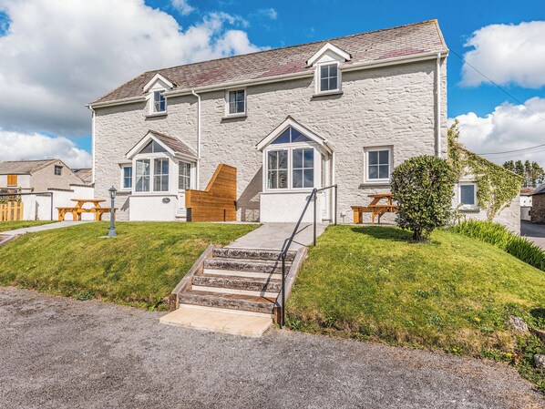 Une maison en pierre à deux étages, avec un escalier en pierre menant à la porte d’entrée, un petit jardin devant la maison agrémenté d’un banc en bois et une allée pavée.