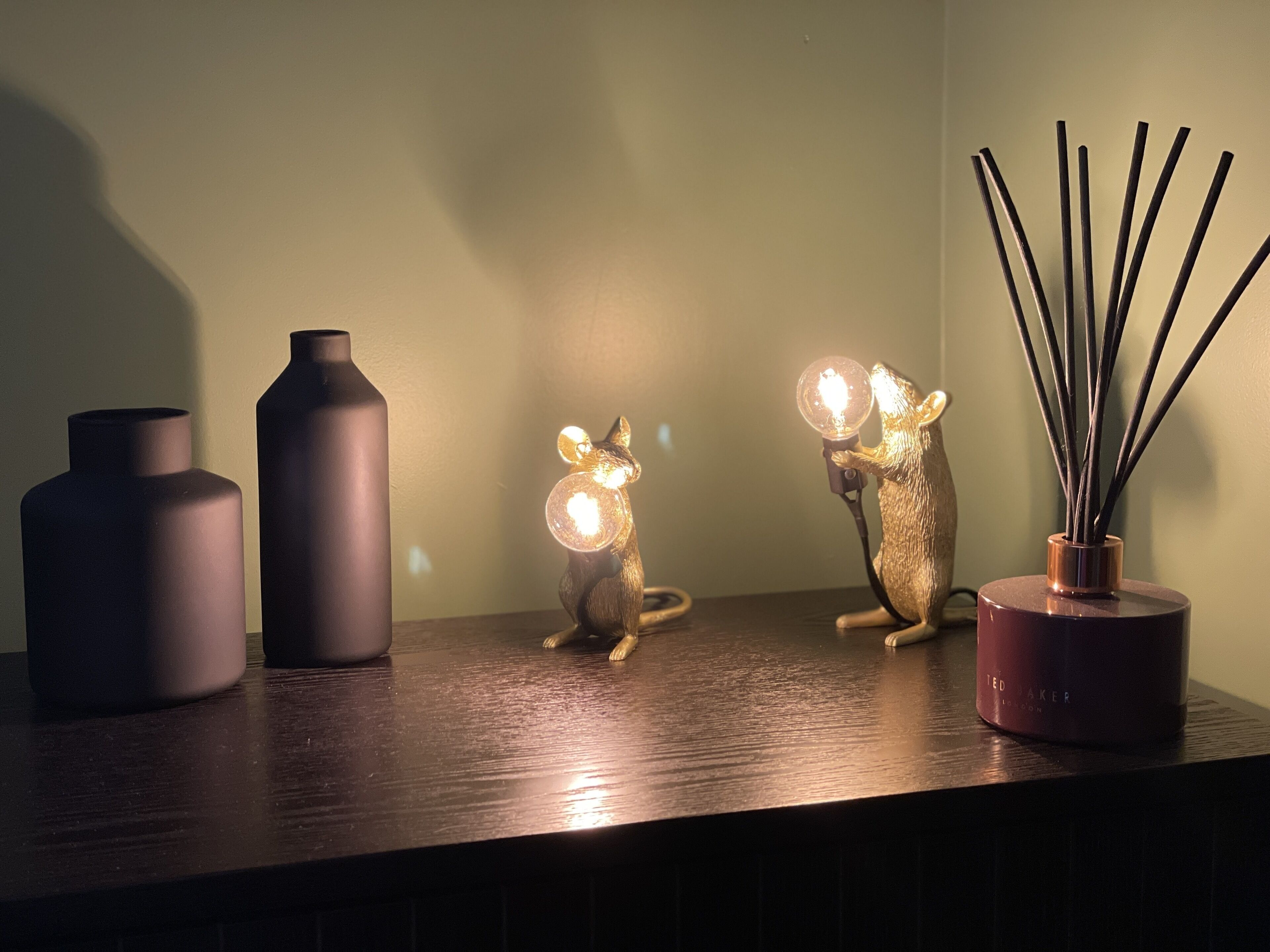 Three decorative items on a wooden surface: two vases and a reed diffuser with a mouse figurine.