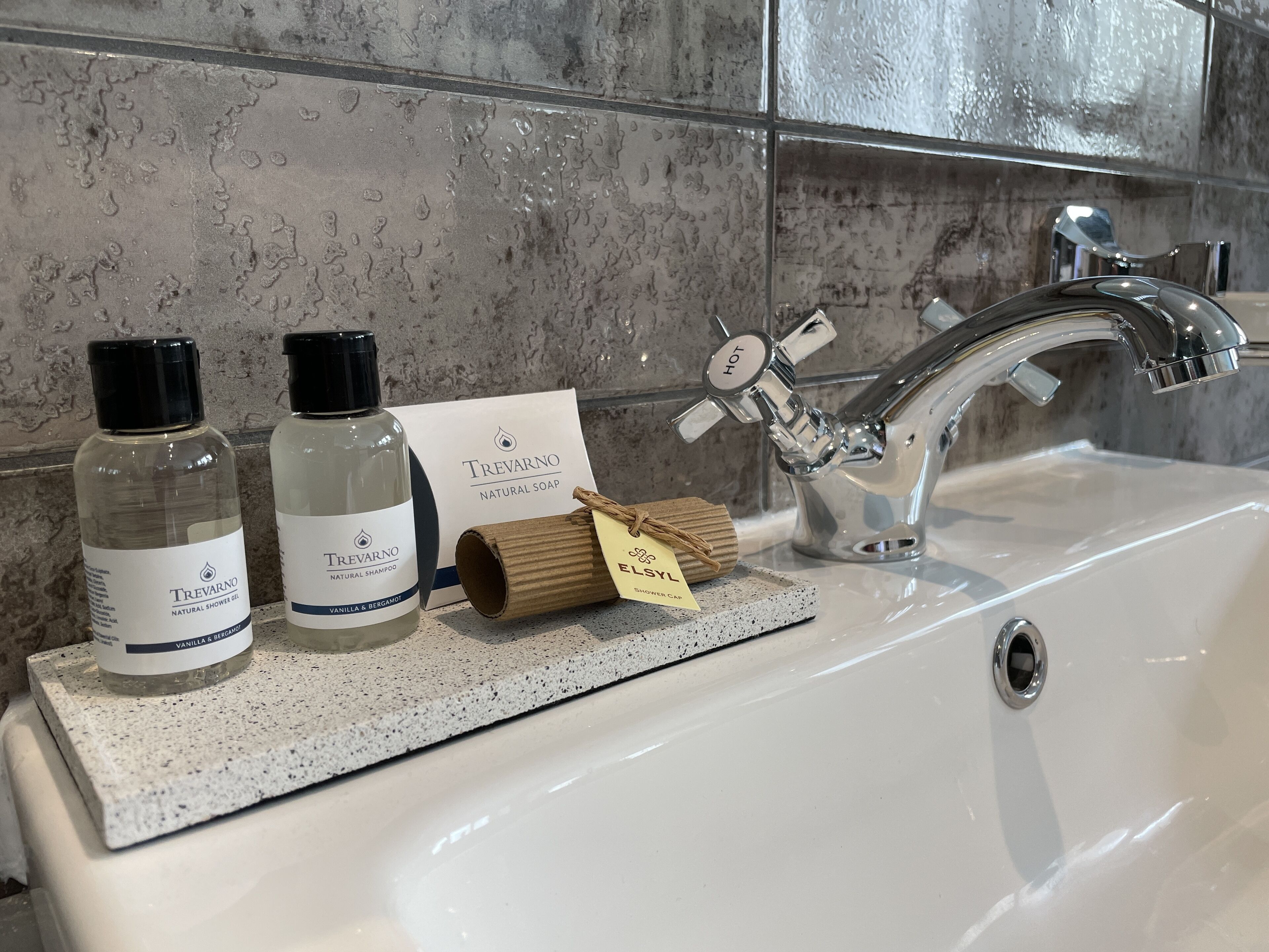 A bathroom sink with two bottles of Trevarno natural shower gel and shampoo, a shower cap, and a "hot" water faucet.