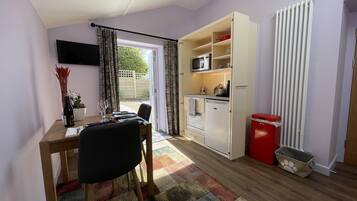 Cocina compacta con microondas, horno y un refrigerador pequeño. Hay una zona de comedor con una mesa y sillas, y una ventana con cortinas. Se observa un radiador en el lado derecho.