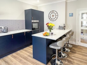 A modern kitchen with dark cabinetry, a white countertop, a built-in oven, a wall clock, and a bar area with stools. - 2 bedroom accommodation in Oban (Oban)