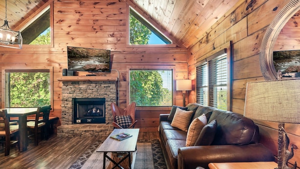 L’intérieur chaleureux d’un chalet avec une cheminée en pierre, un canapé en cuir, un plancher en bois et de grandes fenêtres offrant une vue sur le paysage extérieur.