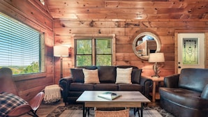 L’intérieur chaleureux d’un chalet avec un plafond en bois, un canapé en cuir marron, un fauteuil assorti, une petite table avec un livre et un panier en osier.