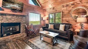 L’intérieur chaleureux d’un chalet avec une cheminée en pierre, un canapé en cuir et un téléviseur fixé au mur.