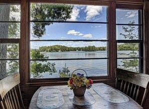 Une table en bois, sur laquelle sont posés un panier tressé rempli de fleurs et un set de table, le tout placé devant une fenêtre donnant sur un lac et des arbres.