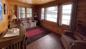 A cozy cabin interior with a plaid sofa, a wooden table with a tray of mugs, and large windows offering a view of the outdoors.