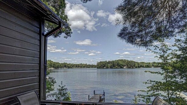 A wooden cabin with a deck overlooking a lake, featuring two chairs and a table.