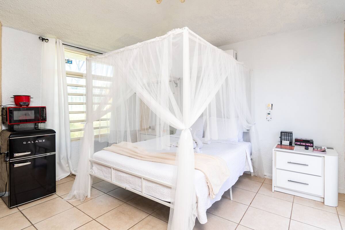 A bedroom with a four-poster bed, a white bedside table, a small black refrigerator, and a red pot on top.