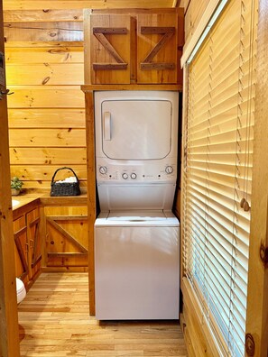 A compact laundry room with a built-in washer and dryer, wooden cabinetry, and a window with blinds. - Big South Fork! Peaceful, Relaxing, On the Trails…Six Decks ~ Nature’s Paradise! (Strunk)