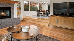 A modern living room with a fireplace, a TV, wooden flooring, and a dining area with a view of the outdoors.