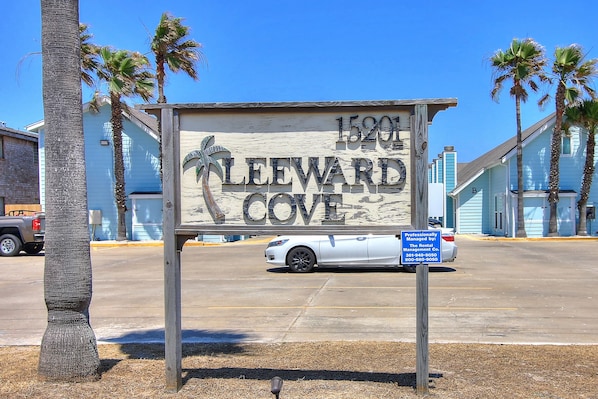 Un panneau en bois portant l’inscription « CLEVARD COVE » et le logo d’un palmier, une voiture blanche et un panneau bleu indiquant qu’il s’agit d’une voiture de location.