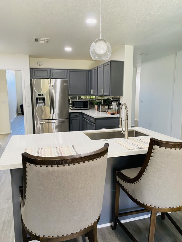 A modern kitchen with a stainless steel refrigerator, a microwave, and a sink. There are two chairs with wooden frames and fabric seats.