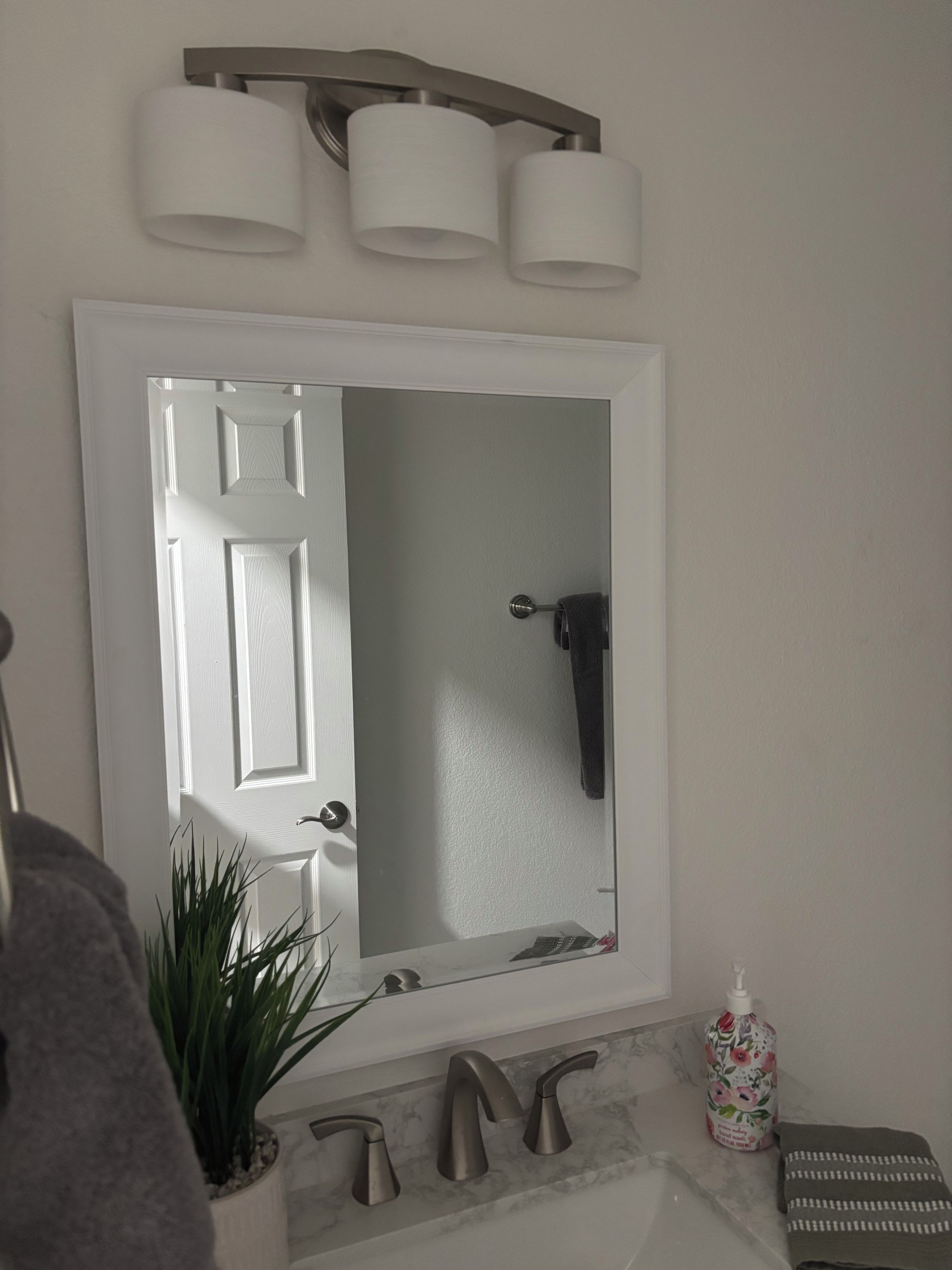 A bathroom with a white door, a mirror, a towel rack, a sink with a marble countertop, and a plant.