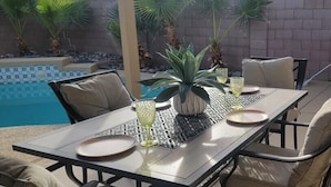 En uteplats med ett bord för fyra personer, omgiven av stolar, en krukväxt och en pool i bakgrunden.