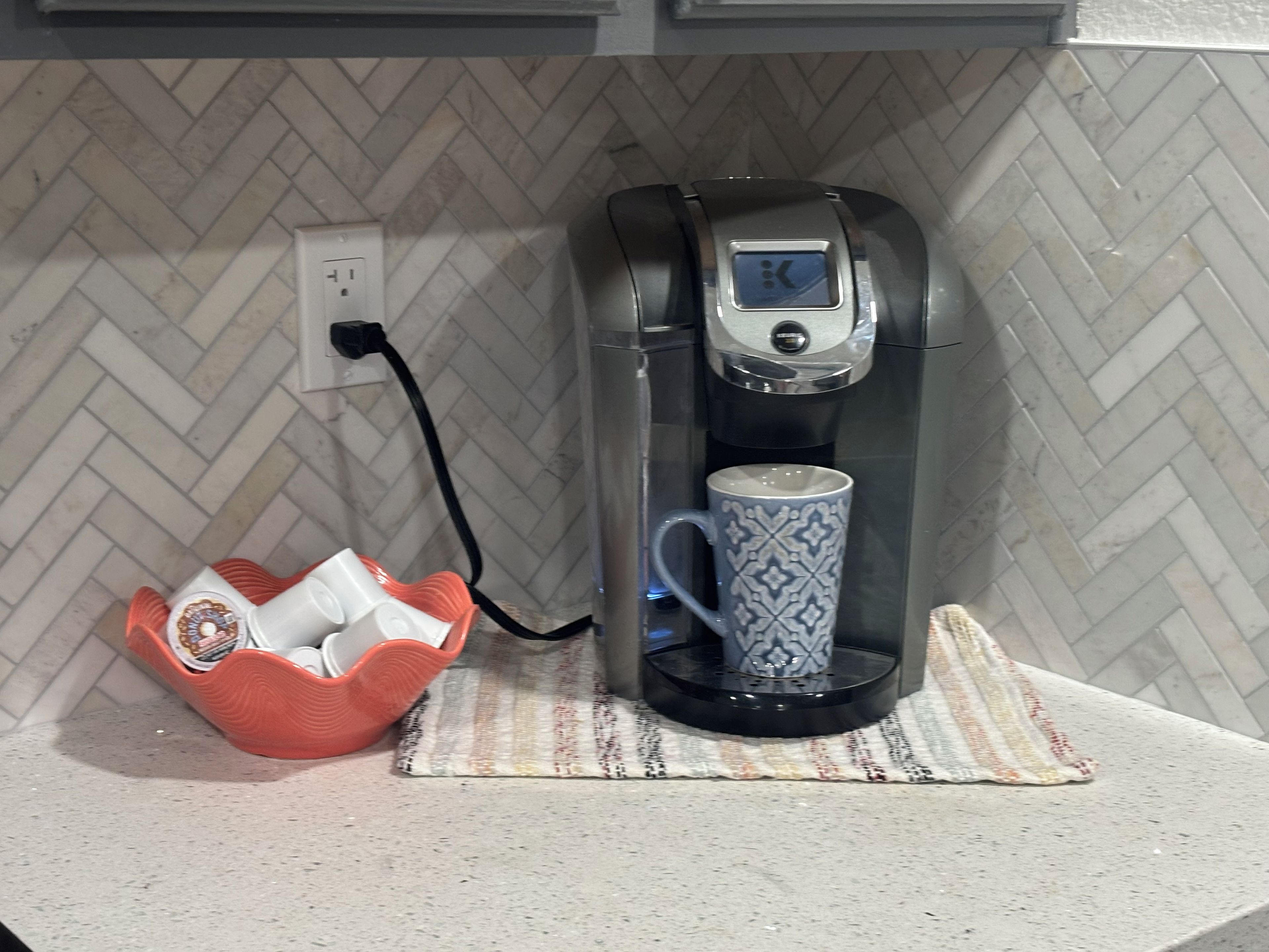 A coffee maker on a kitchen counter with a power outlet and a basket of coffee pods.