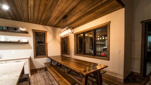 Een keuken met houten werkbladen, een grote houten tafel en inbouwbanken. Er zijn ramen met gordijnen, een hanglamp en planken met keukengerei.