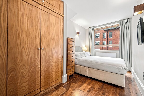 A bedroom with a large wooden wardrobe, a bed with a headboard, and a window with curtains.