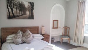 A bedroom with a wooden bed, white bedding, a painting of trees, a mirror, and a chair.