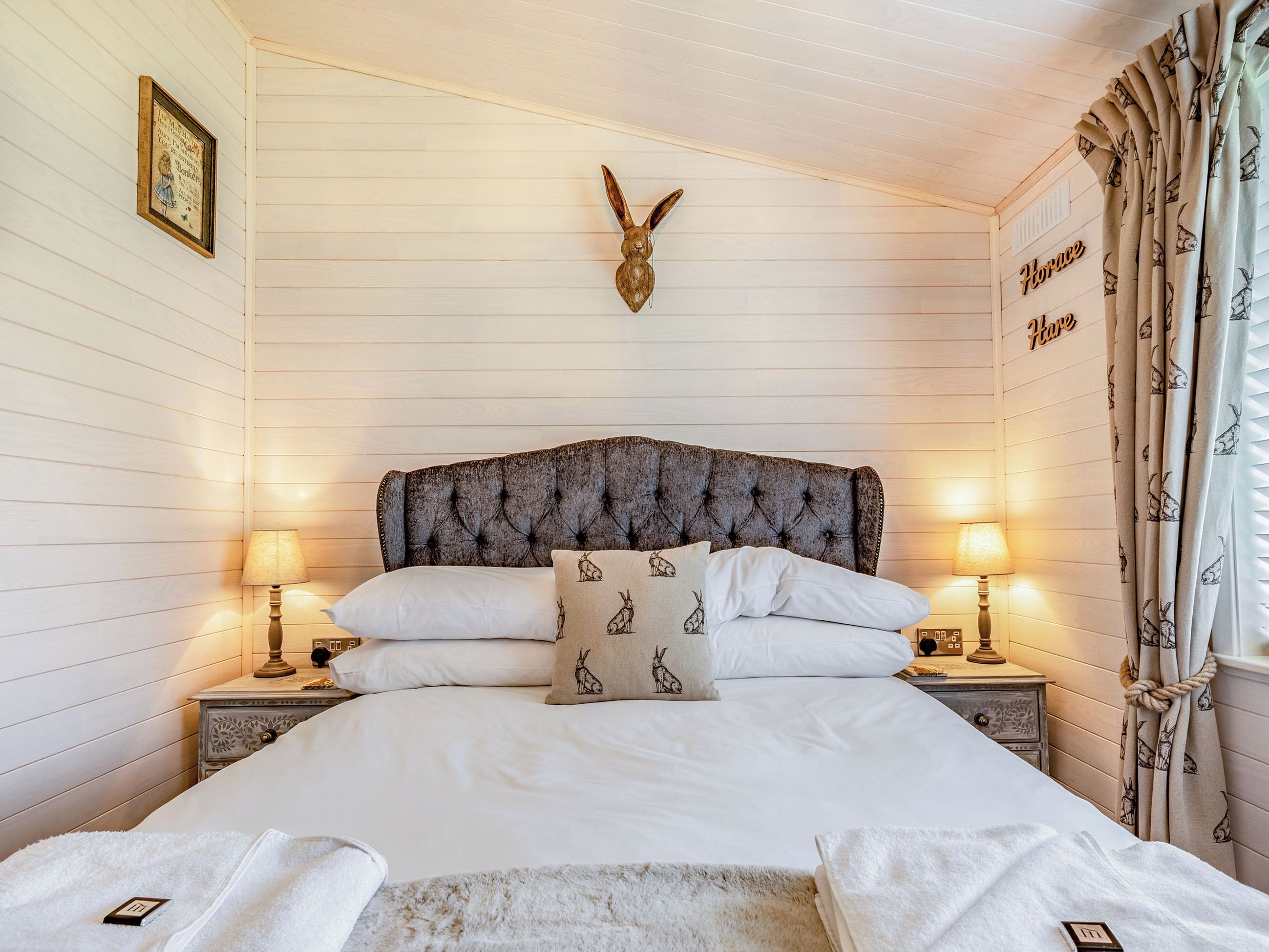 Une chambre à coucher avec une tête de lit capitonnée, du linge de lit blanc, des coussins décoratifs, des lampes de chevet, un tableau encadré et un panneau décoratif sur le mur.
