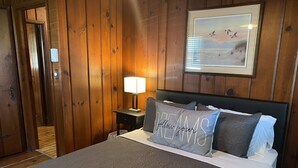 Une pièce aux murs lambrissés, comprenant un lit, une table de chevet, un tableau encadré et une porte.