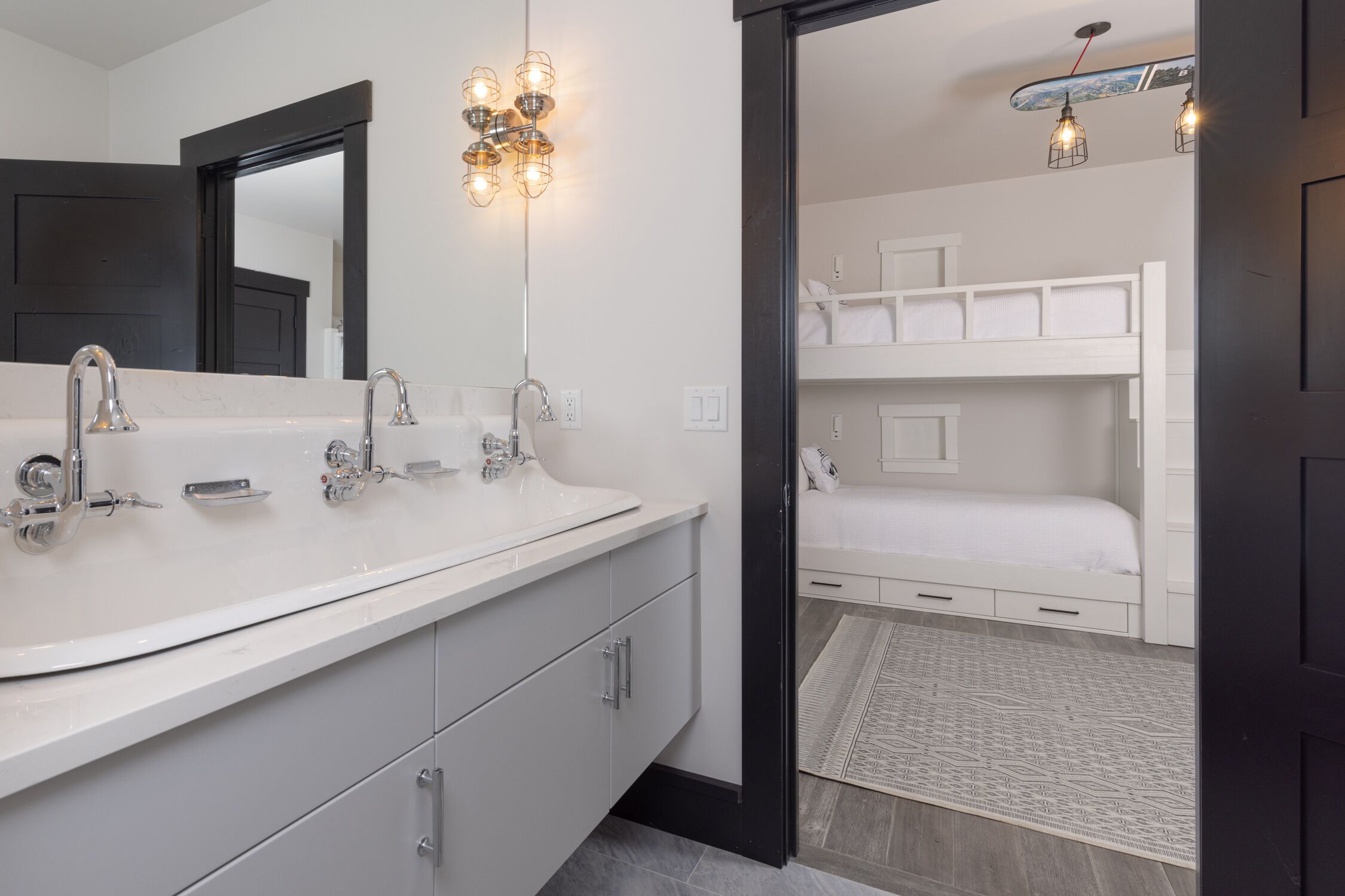 Un baño moderno con un lavamanos grande blanco, dos grifos y un espejo. Se observa una cama blanca con cabecera y una pequeña alfombra en el suelo.