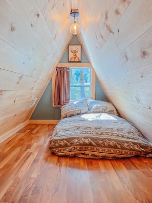 Une chambre mansardée chaleureuse avec un plafond en pente, un lit recouvert d’une literie à motifs, une fenêtre donnant sur les arbres et un tableau représentant un cerf, accroché au mur.