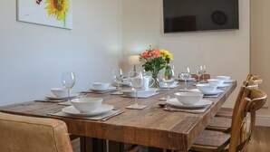 Une salle à manger avec une longue table en bois dressée pour le repas, un vase rempli de fleurs et un téléviseur à écran plat fixé au mur.