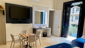 Habitación de hotel con cama, escritorio con silla, televisor montado en la pared, baño con lavamanos y balcón con vista a edificios.