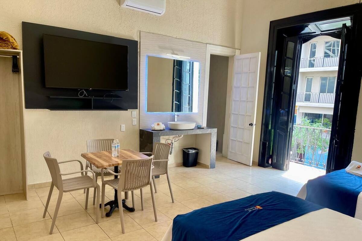 A hotel room with a bed, a desk with a chair, a TV mounted on the wall, a bathroom with a sink, and a balcony with a view of buildings.