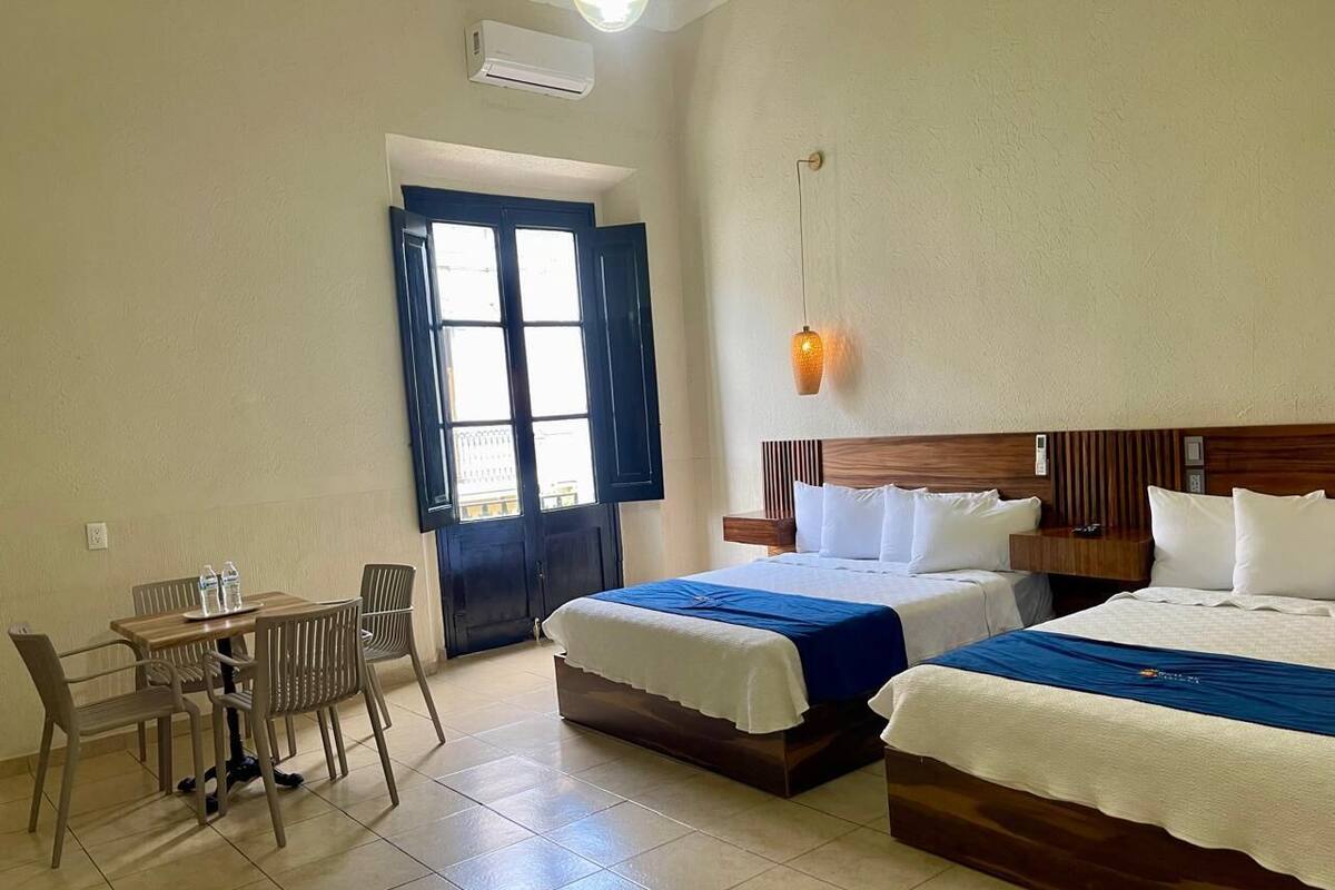 A hotel room with two beds, a dining table with chairs, and a large window with a view.