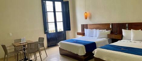 A hotel room with two beds, a dining table with chairs, and a large window with a view.